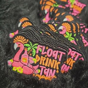 May include: A stack of embroidered patches featuring a pink flamingo float, a green palm tree, and a bottle. The text reads FLOAT DRINK TAN repeat in pink and orange lettering against a black background with a black stitched border.