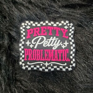 May include: A square embroidered patch with rounded corners featuring a black background and a white and black checkered border. The text PRETTY in pink, Petty in white script, and PROBLEMATIC in pink is centered with small white star accents.