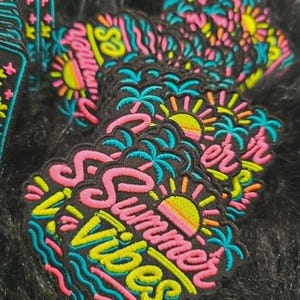 May include: A collection of embroidered patches featuring a black background with the text Summer Vibes in pink and yellow. The design includes a yellow and pink sun, blue palm trees, and wavy blue lines, all outlined in a thick black border.