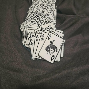 May include: A collection of white rectangular patches shaped like playing cards, each featuring a black embroidered letter A and a suit symbol. One prominent patch displays a black embroidered graphic of a bucking bronco and rider.
