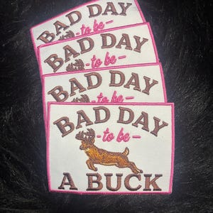 May include: Four rectangular embroidered patches with a white background and pink border. The text reads BAD DAY to be A BUCK in brown and pink thread. A brown deer illustration is positioned between the words to be and A BUCK.