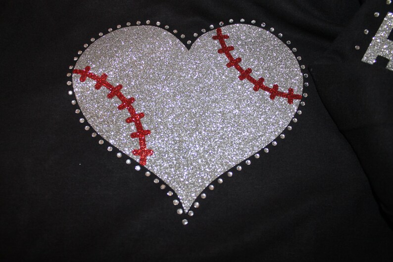 May include: A silver glitter heart with a red baseball stitch design. The heart is outlined with silver rhinestones.