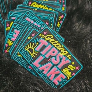 May include: A rectangular embroidered patch with a black background featuring the yellow text Getting Tipsy at the, pink text LAKE, a yellow sun, blue wavy lines representing water, and small pink cross symbols scattered across the design.