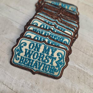 May include: A stack of rectangular embroidered patches with scalloped edges. Each patch features a beige background with a dark brown rope-style border and teal text that reads ON MY WORST BEHAVIOR, accented by decorative teal scrollwork designs.