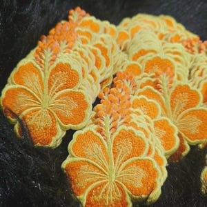 May include: A collection of embroidered hibiscus flower patches featuring vibrant orange petals with detailed yellow stitching along the edges and a prominent central stamen. Each patch displays a consistent floral shape and color scheme.