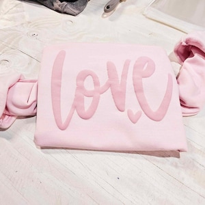 May include: A pink sweatshirt with the word "love" written in a large, cursive font. The sweatshirt is folded in half and the sleeves are rolled up.