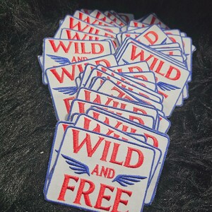 May include: A stack of rectangular embroidered patches with a white background, a blue border, and red text that reads WILD AND FREE. Each patch features a small blue wing graphic on either side of the word AND, stitched in a bold, clear font.