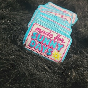 May include: A stack of rectangular embroidered patches with a white background and a light blue border. The text reads made for SUNNY DAYS in pink and blue letters, accompanied by a small yellow sun icon stitched in the bottom right corner.