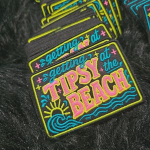 May include: A rectangular embroidered patch with a black background and a yellow border. The text reads getting at the TIPSY BEACH in pink and blue. A yellow sun and blue ocean waves are embroidered at the bottom with small pink and yellow stars.