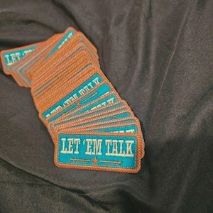 May include: A stack of rectangular embroidered patches with a teal background and a copper-colored border. Each patch features the text LET 'EM TALK in white lettering, accompanied by a small star and horizontal lines centered below the text.