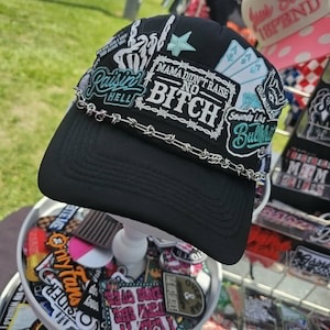 May include: A black baseball cap featuring a silver-toned chain across the brim and multiple embroidered patches. The patches include text reading "MAMA DIDN'T RAISE NO BITCH," "Raisin HELL," and "Sounds Like Bullshit," alongside playing card graphics.