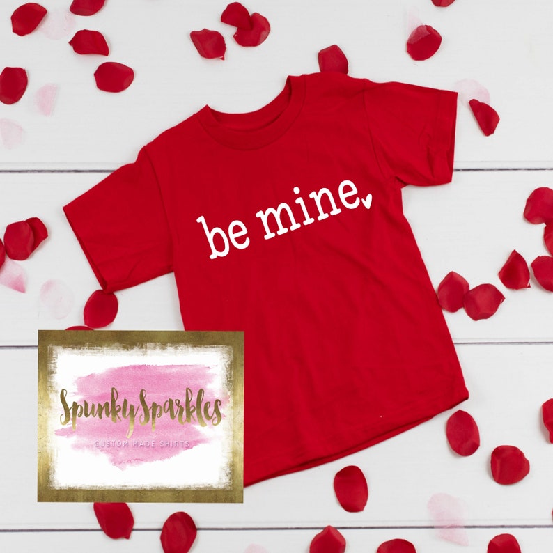 Może przedstawiać: Czerwona koszulka z napisem "be mine" w białych literach. Koszulka jest otoczona czerwonymi płatkami r&oacute;ż na białym tle. Obraz zawiera r&oacute;wnież logo "Spunky Sparkles Custom Made Shirts".