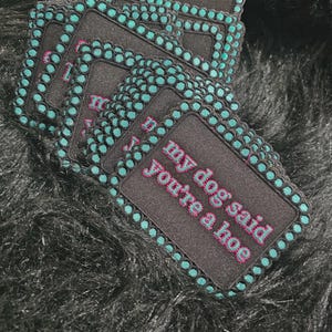 May include: A stack of rectangular black embroidered patches with a teal dotted border. Each patch features the text "my dog said you're a hoe" stitched in pink thread with a teal outline. The patches have a structured, fabric-based design.