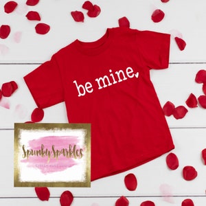 Może przedstawiać: Czerwona koszulka z napisem "be mine" w białych literach. Koszulka jest otoczona czerwonymi płatkami r&oacute;ż na białym tle. Obraz zawiera r&oacute;wnież logo "Spunky Sparkles Custom Made Shirts".