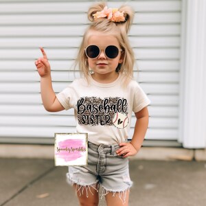 Könnte beinhalten: Ein junges Mädchen trägt ein weißes T-Shirt mit Leopardenmuster und dem Text "Baseball Sister" mit einem Baseball-Motiv. Sie trägt Jeans-Shorts und eine Sonnenbrille.
