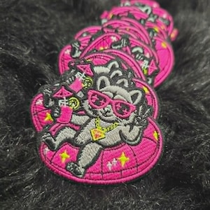 May include: A stack of embroidered patches featuring a gray cat wearing bright pink sunglasses and a gold-colored diamond pendant necklace. The cat is lounging on a pink inflatable ring, holding a pink cocktail with a small umbrella and lime slice.