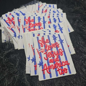 May include: A rectangular embroidered patch featuring a white background with vertical blue stripes and blue stars. The red embroidered text reads She loves Jesus and America too, accompanied by a small embroidered American flag icon.