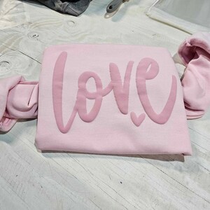 May include: Pink sweatshirt with the word "LOVE" in a pink, cursive font.