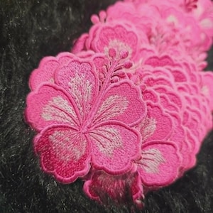 May include: A collection of bright pink hibiscus flower embroidered patches. Each patch features detailed stitching with a prominent central stamen and scalloped petal edges, layered together to show the intricate thread work and vibrant color.