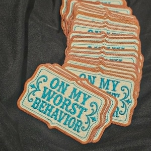May include: A stack of rectangular embroidered patches with scalloped edges. Each patch features a cream-colored background with a copper-colored border and teal-colored text that reads ON MY WORST BEHAVIOR, accented by decorative scrollwork.