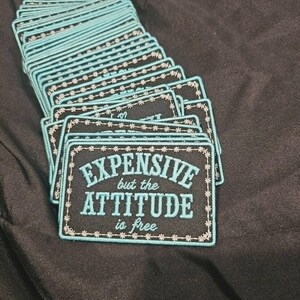 May include: A stack of rectangular embroidered patches featuring a black background with light blue text that reads EXPENSIVE but the ATTITUDE is free. Each patch has a light blue border with a small star pattern stitched along the edges.