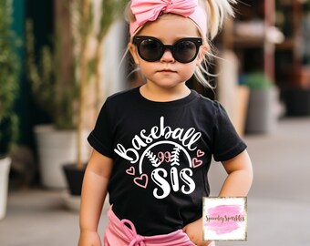 Puede incluir: Una camiseta negra con un gráfico blanco de una pelota de béisbol con el texto "Baseball Sis" y corazones rosas. La niña lleva pantalones rosas y una diadema rosa con un lazo.