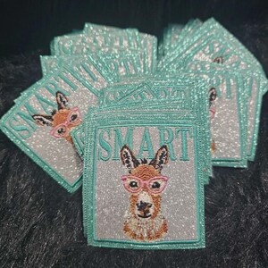 May include: A stack of square embroidered patches featuring a brown donkey wearing pink glasses. The background of each patch is silver glitter fabric with a teal border. The word SMART is embroidered in teal letters above the donkey's head.