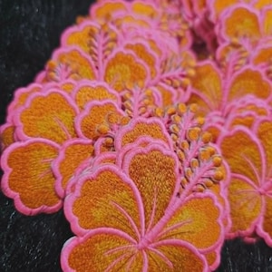 May include: A collection of embroidered hibiscus flower patches featuring vibrant orange petals with detailed golden-yellow centers, each outlined with a bright pink stitched border and a prominent stamen extending from the side of the bloom.