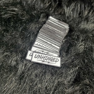 May include: A stack of rectangular embroidered patches with a white background and black border. Each patch features the word UNHINGED in a stylized black font with jagged, lightning-like lines extending from the letters across the surface.