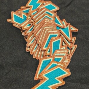May include: A collection of multiple lightning bolt patches, each featuring a bright teal center surrounded by a thick, textured copper-colored embroidered border. The patches are stacked in a loose pile, showing the consistent shape and color.