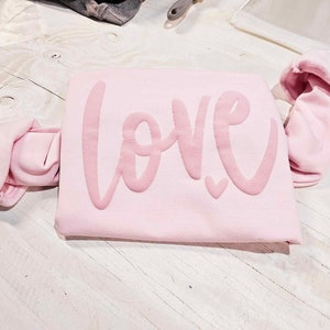 May include: A pink sweatshirt with the word "love" written in a large, cursive font. The sweatshirt is folded in half and the sleeves are rolled up.