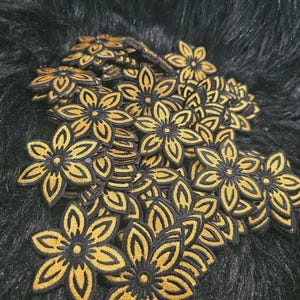 May include: A collection of multiple embroidered flower patches, each featuring a six-petaled design with a golden yellow center and thick black outlines. The patches are layered together, showcasing the consistent floral shape and color scheme.