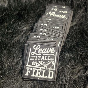 May include: A stack of square black embroidered patches featuring white text that reads Leave IT ALL on the FIELD, accompanied by a graphic of a football goal post and a football. The patches have a black border and a textured fabric finish.