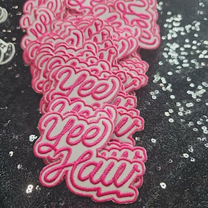 May include: A stack of embroidered patches featuring the text Yee Haw in a bright pink cursive font with a white background and a pink border. The patches are layered on top of each other, showcasing the repeating design and the stitched texture.
