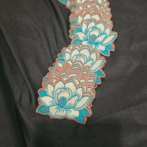 May include: A stack of embroidered lotus flower patches featuring cream-colored petals with vibrant teal blue accents and a copper-colored border. The floral design is repeated across multiple identical fabric patches layered vertically.