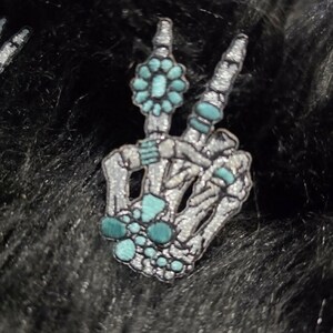 May include: A silver-toned skeleton hand patch making a peace sign gesture, adorned with turquoise-colored accents including a flower shape on the index finger, stacked rings on the fingers, and a cluster of circular stones at the wrist base.