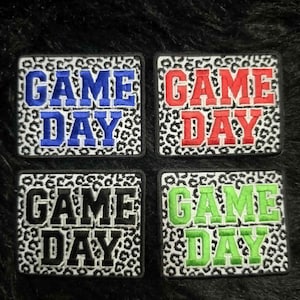 May include: Four rectangular embroidered patches featuring a white and black leopard print background. Each patch displays the text GAME DAY in bold block letters, with one patch in blue, one in red, one in black, and one in bright lime green.