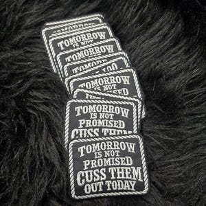 May include: A stack of rectangular black embroidered patches with a white rope-style border. Each patch features the white text: TOMORROW IS NOT PROMISED CUSS THEM OUT TODAY. The design is consistent across all stacked pieces, showing clear white lettering.