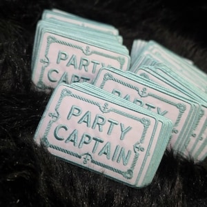 May include: A stack of rectangular embroidered patches featuring a light blue background with a dark teal border resembling a rope. The text PARTY CAPTAIN is embroidered in dark teal in the center, with small anchor icons at the top and bottom.