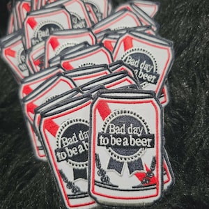 May include: A collection of embroidered patches shaped like beer cans. Each patch features a white background with red accents and a central black circular emblem containing the text Bad day to be a beer in white lettering with wheat stalks.