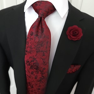 Puede incluir: Un traje negro con camisa blanca, corbata burdeos con estampado floral, pañuelo a juego y un prendedor de solapa de rosa burdeos. La corbata y el pañuelo presentan un diseño floral oscuro.