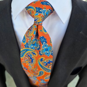 May include: A necktie featuring a vibrant orange background with a detailed paisley pattern in shades of bright blue, navy, and yellow. The tie is knotted and worn over a crisp white collared shirt, which is layered under a dark black suit jacket.