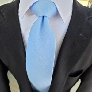 Peut inclure: Une cravate bleu clair avec une texture tissée visible, nouée de manière classique. La cravate est portée sur une chemise blanche à col rigide, superposée sous un blazer noir uni. Le tissu de la cravate semble mat et de couleur uniforme.