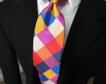Könnte beinhalten: Eine Krawatte mit einem lebendigen, mehrfarbigen Rautenmuster in den Farben Magenta, Königsblau, Goldgelb, Orange, Rot und Creme. Der Stoff hat eine feine, diagonal gerippte Textur und wird zu einem weißen Hemd und einem schwarzen Blazer getragen.