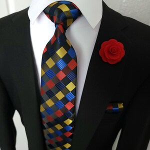 Puede incluir: Un traje negro con una camisa de cuello blanco, una corbata estampada, una flor de solapa roja y un pañuelo de bolsillo a juego. La corbata presenta un patrón repetitivo de cuadrados rojos, amarillos, azules y negros.