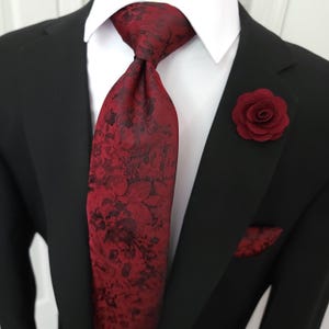 May include: A dark red necktie featuring a black floral pattern, paired with a matching dark red pocket square and a dark red fabric rose lapel pin. The set is worn over a crisp white collared shirt and a solid black blazer jacket.