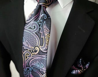 May include: A necktie featuring a dark gray base with an intricate paisley pattern in shades of light blue, pink, and gold. The tie is worn over a crisp white collared shirt and a black blazer, with a matching pocket square visible in the jacket pocket.