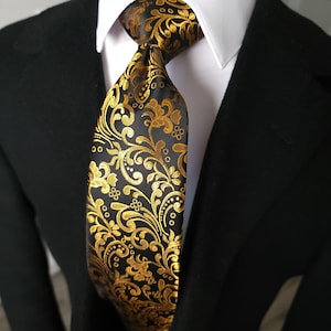 Op de afbeelding: Een stropdas met een zwarte basis en een metallic goudkleurig bloemen- en paisley-patroon. De das is geknoopt en gedragen over een wit overhemd met kraag, dat onder een effen zwart colbertjasje zit, wat zorgt voor een formele uitstraling.