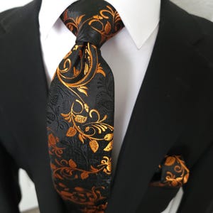 Puede incluir: Una corbata negra con un diseño floral naranja, combinada con un pañuelo de bolsillo a juego. La corbata está anudada y se lleva con una camisa blanca y una chaqueta de traje negra. El diseño floral es detallado y ornamentado.