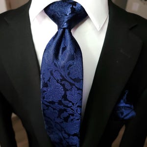 May include: A dark blue necktie featuring a tonal floral and paisley jacquard pattern, worn with a crisp white collared shirt, a black suit jacket, and a matching dark blue pocket square tucked into the jacket breast pocket.